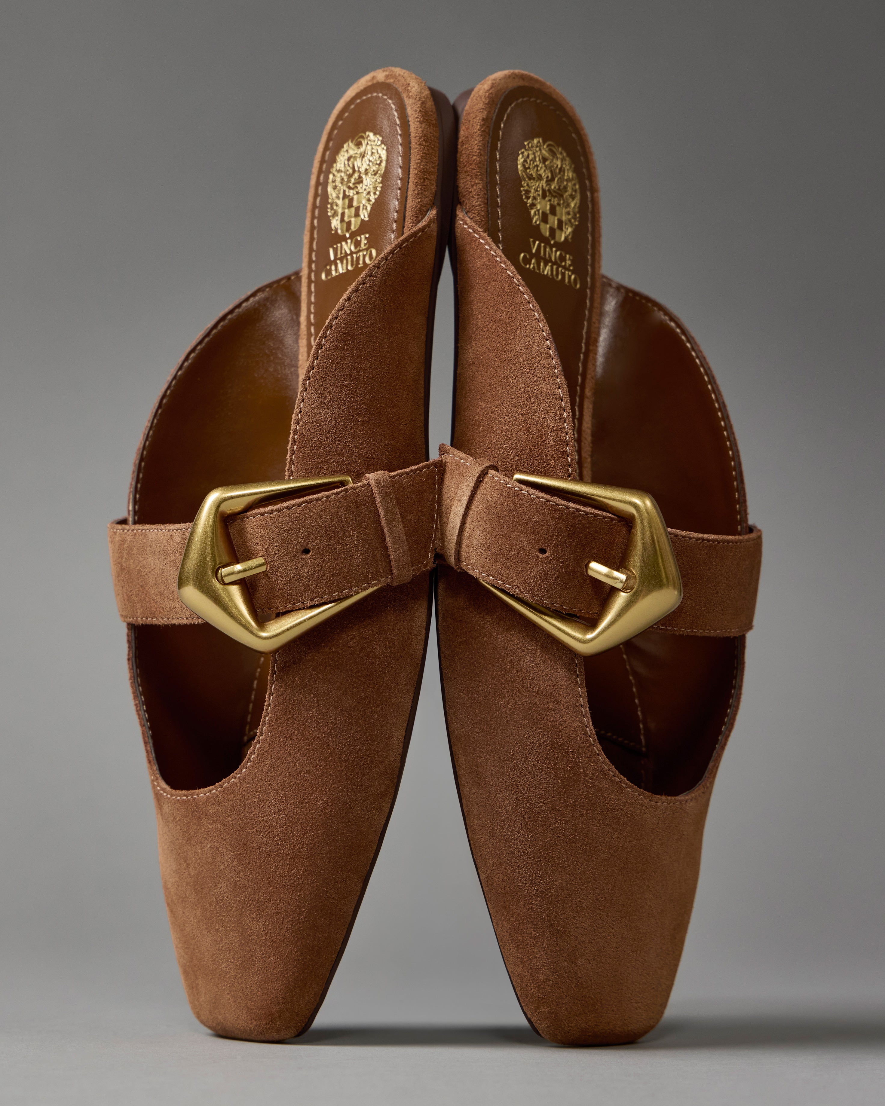 The Vellma Buckle Mule by Vince Camuto features brown suede leather, oversized gold buckles, pointed toes, tan leather insoles with branding, and a slip-on design for effortless style and comfort—all shown against a plain gray background.