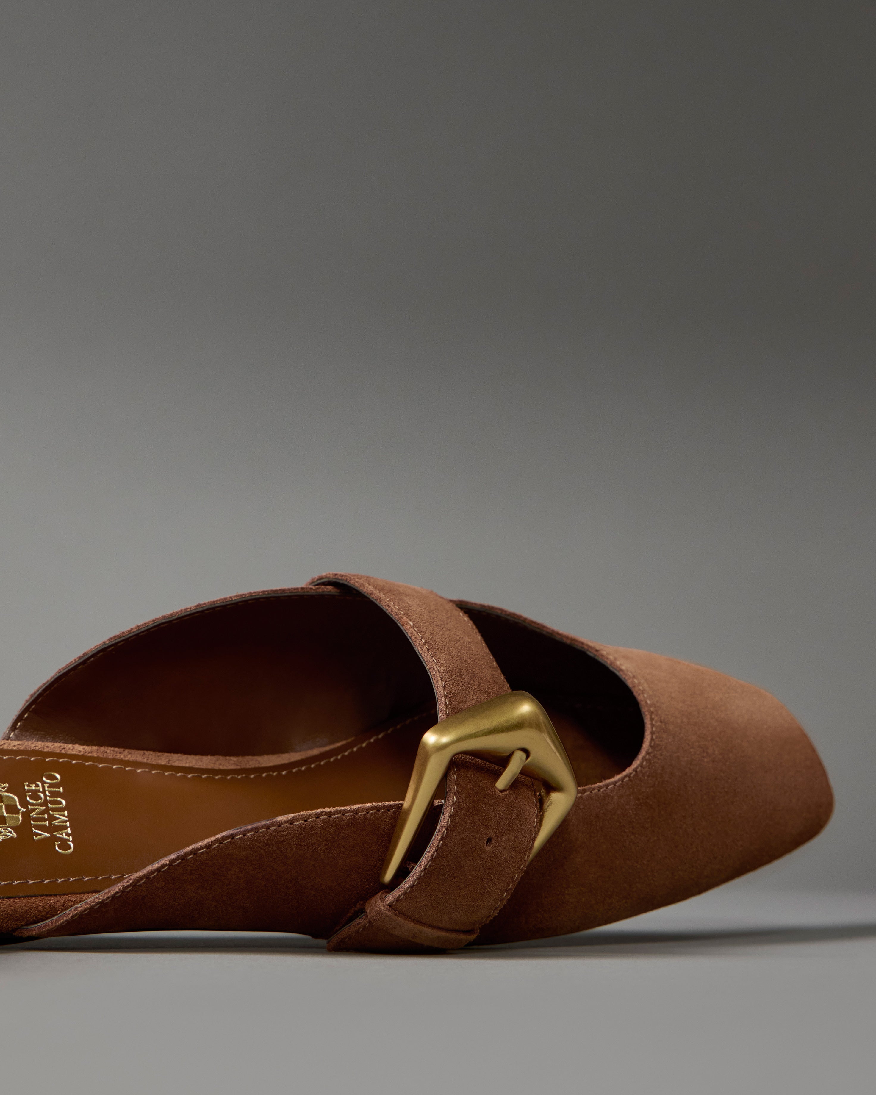 A close-up of the Vince Camuto Vellma Buckle Mule, a brown suede pointed-toe slip-on flat with a large gold buckle, shown on a plain gray background.