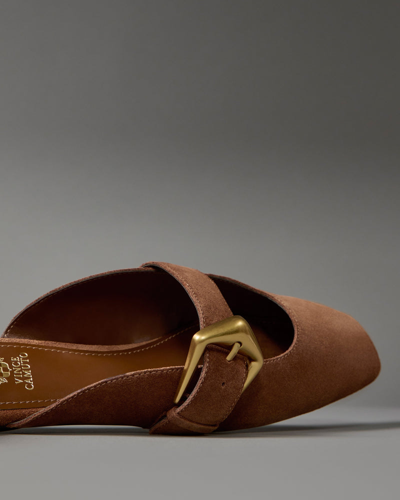 A close-up of the Vince Camuto Vellma Buckle Mule, a brown suede pointed-toe slip-on flat with a large gold buckle, shown on a plain gray background.