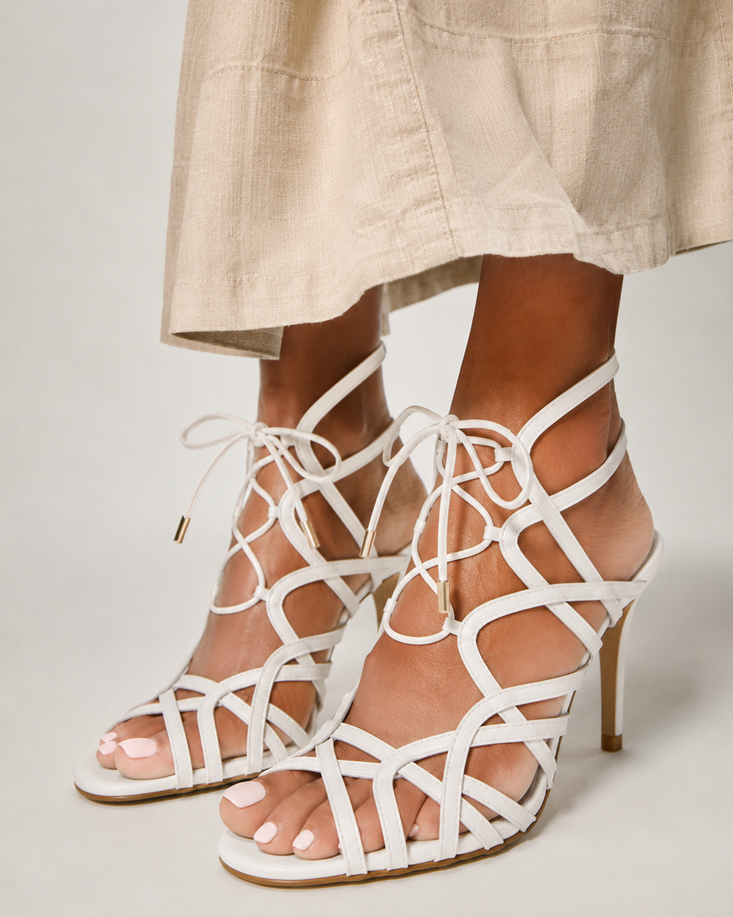 Close-up of a person wearing the Vince Camuto Tilly Sandal—chic white strappy stilettos with geometric cutouts and gold-tipped lace-up design—paired with a beige skirt.