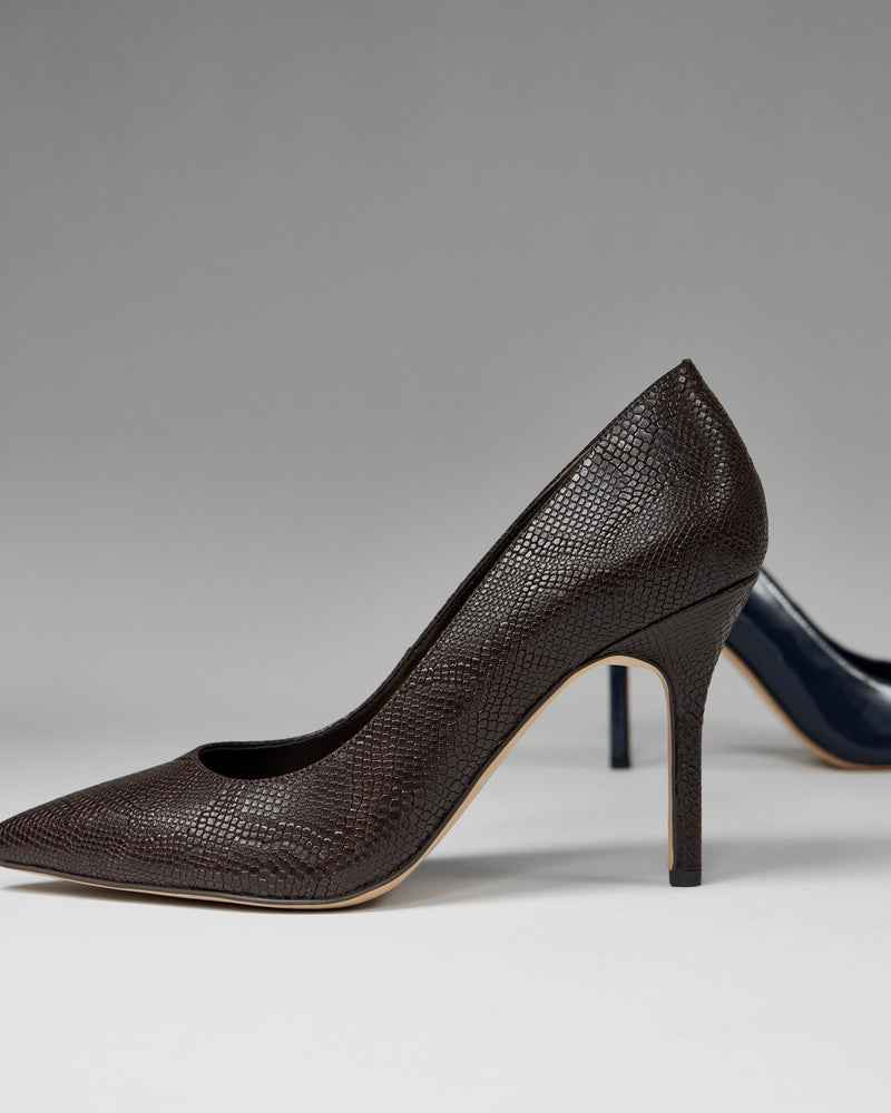 A close-up of the Vince Camuto Maisha Pump, a brown textured high heel with a pointed toe, set against a neutral background. Part of another dark-colored high heel is visible in the background.