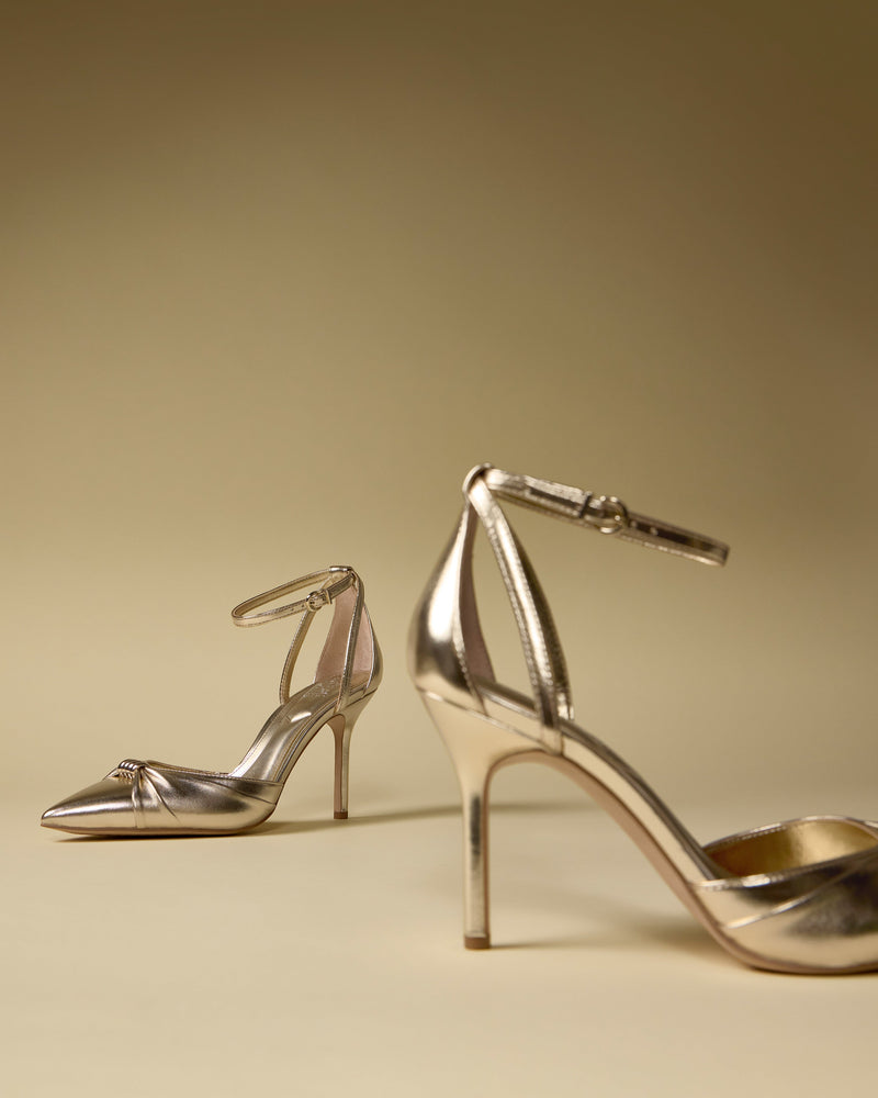 Two Vince Camuto Mallie Pump metallic gold high heels with pointed toes and ankle straps are displayed on a beige surface, one in the foreground and the other slightly behind against a neutral backdrop.