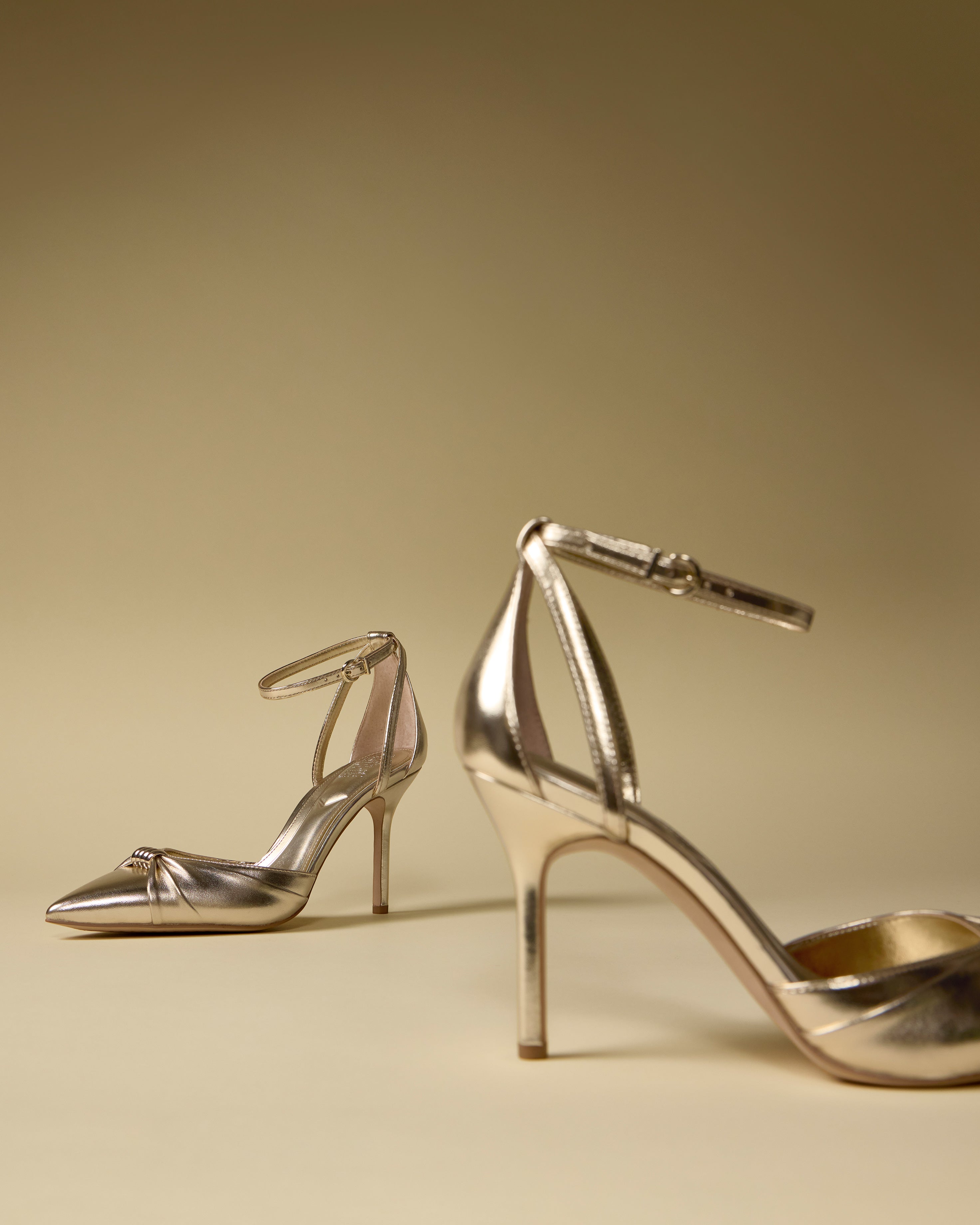 The Vince Camuto Mallie Pump features two elegant, metallic gold high heels with ankle straps displayed on a beige surface and matching background—one in the foreground, the other set farther back.