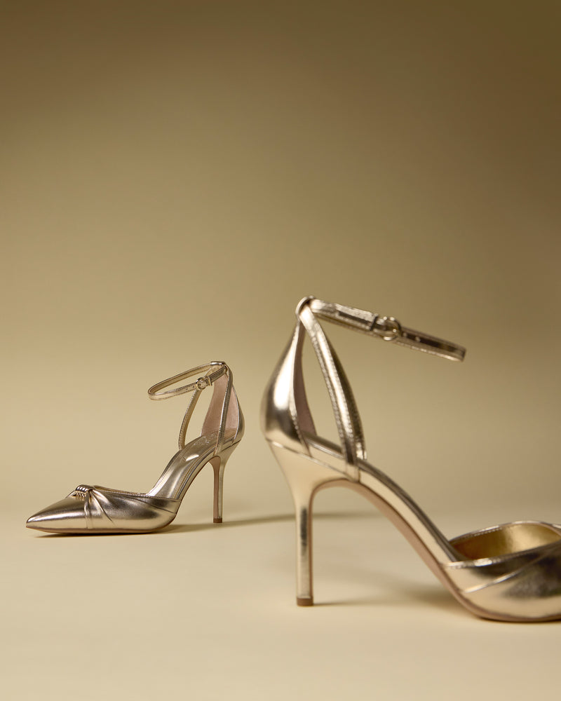 The Vince Camuto Mallie Pump features two elegant, metallic gold high heels with ankle straps displayed on a beige surface and matching background—one in the foreground, the other set farther back.