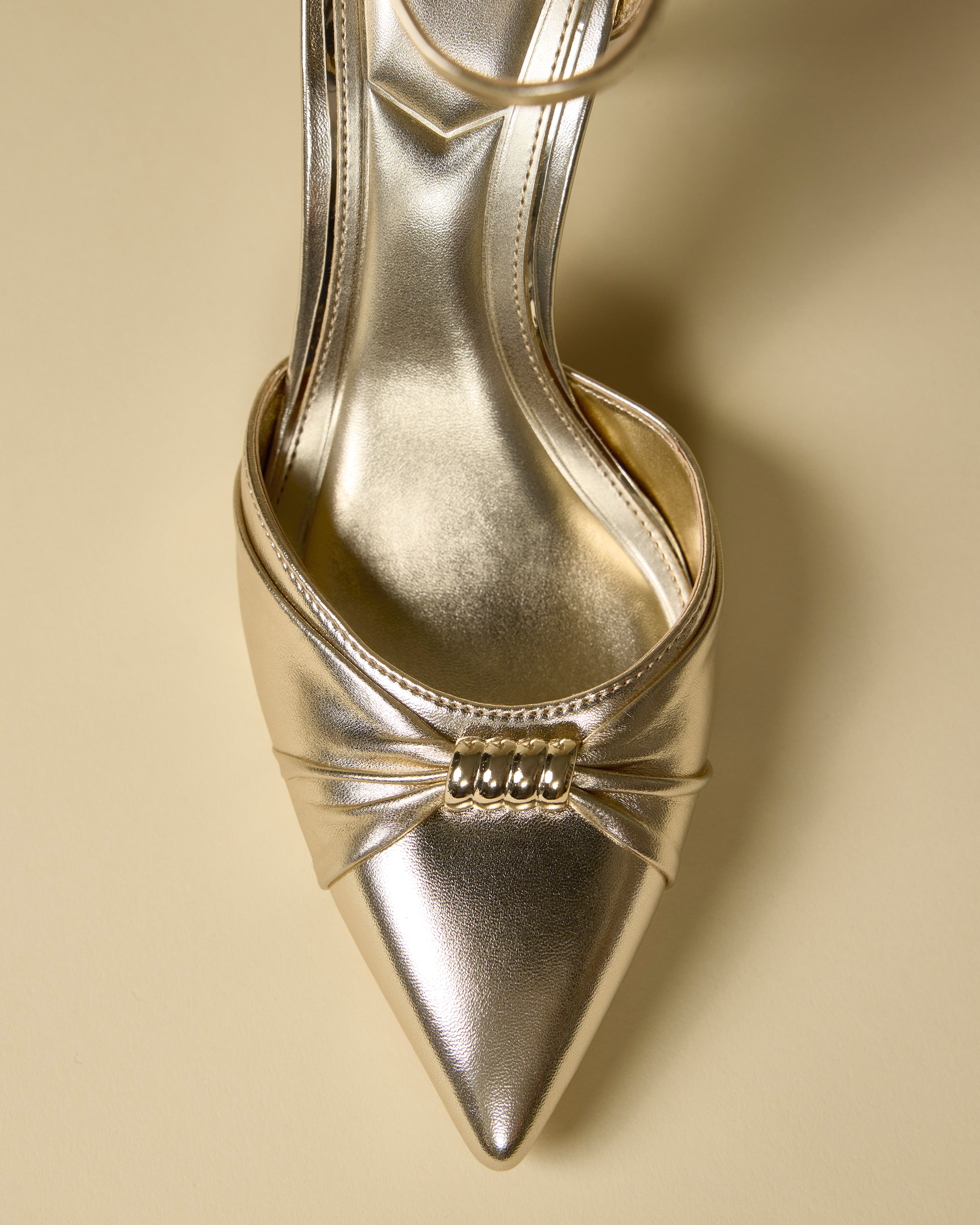 A close-up top view of the Mallie Pump by Vince Camuto, featuring a shiny gold finish, pointed toe, decorative pleats, and a gold embellishment at the front, displayed on a beige background.