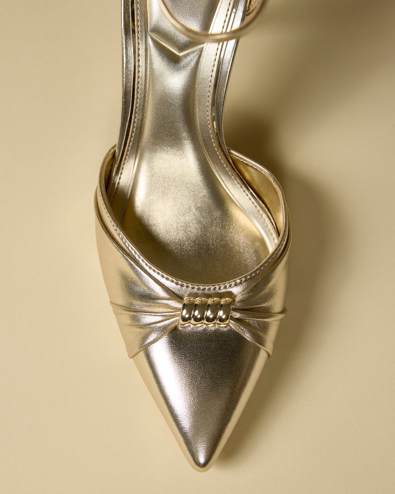 A close-up top view of the Mallie Pump by Vince Camuto, featuring a shiny gold finish, pointed toe, decorative pleats, and a gold embellishment at the front, displayed on a beige background.