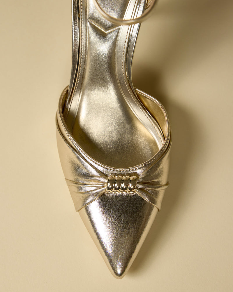 A close-up of the Vince Camuto Mallie Pump shows its gold pointed toe, decorative pleating, and a trio of metallic beads on the front, set against a neutral beige background.