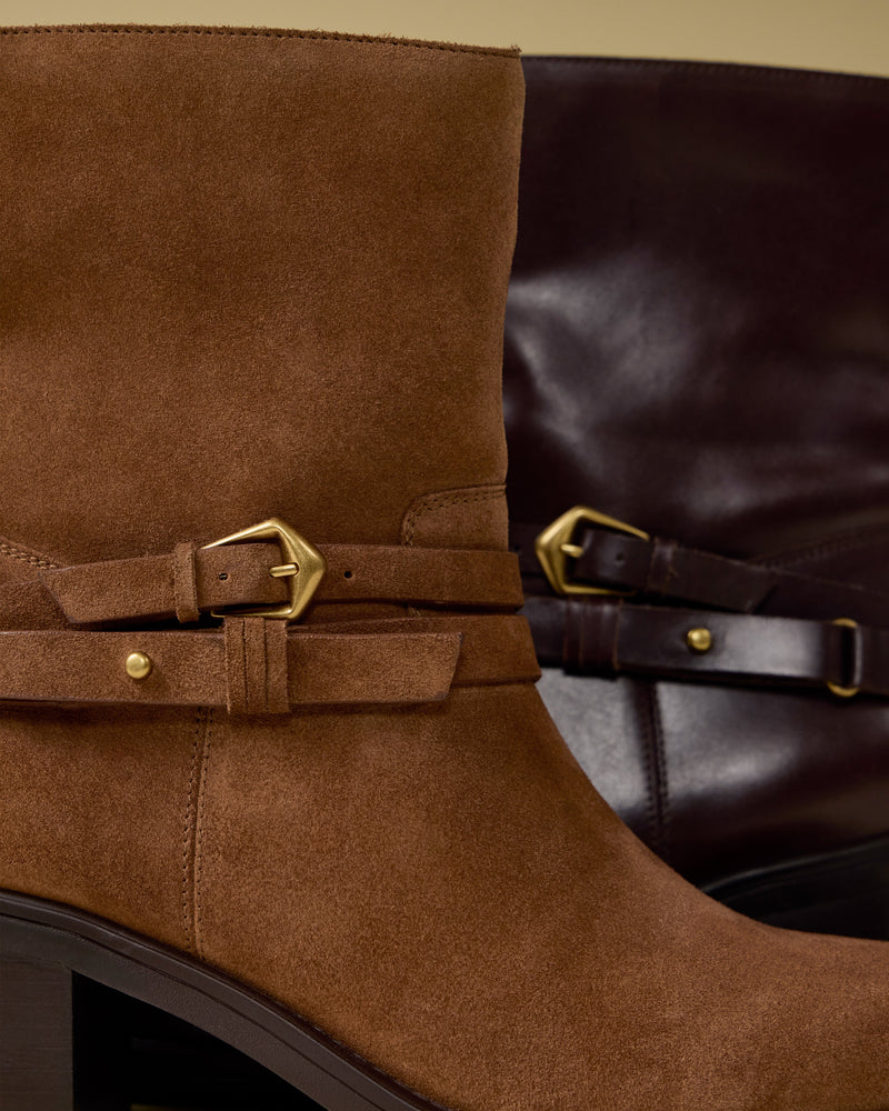 Close-up of two Vince Camuto Armia Booties: one in brown suede with a gold buckle and a stylish harness accent, and the other in dark brown leather with a matching gold buckle. Both boots feature mid-height block heels.