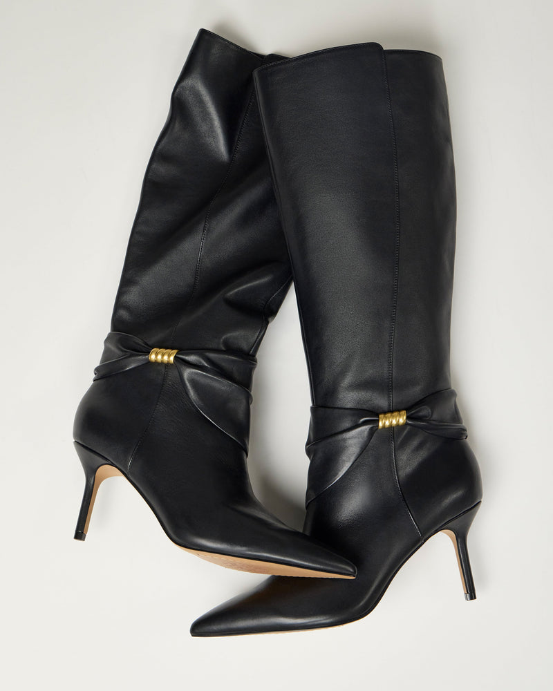 The Meezo Knee High Boot by Vince Camuto features tall black leather, pointed toes, gold bead hardware accents, a skinny heel, and decorative ankle folds, displayed against a light background.
