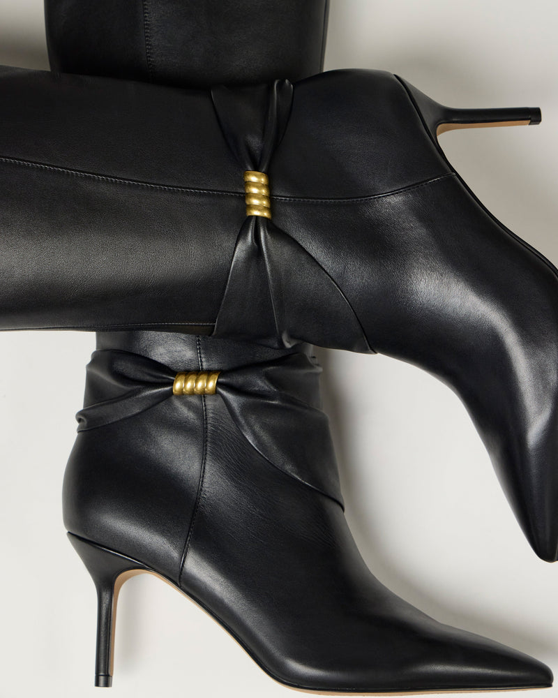 Close-up of Vince Camuto Meezo Extra Wide Calf Knee High Boots in black leather with pointed toes and gold hardware at the gathered ankles. These sleek boots feature skinny heels and are set on a light, neutral background.