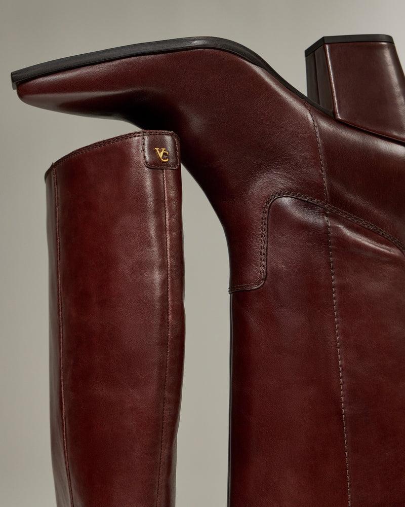 Close-up of the Pamlia Extra Wide Calf Knee High Boot by Vince Camuto, featuring smooth brown leather, a block heel, and a small gold VC logo near the top for subtle western-inspired style.