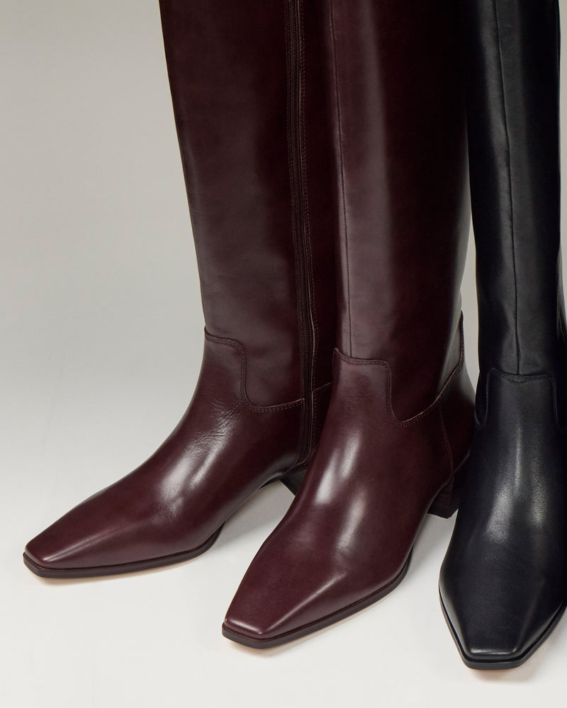 Close-up of two pairs of Vince Camuto Pavla Extra Wide Calf Knee High Boots—one dark burgundy, one black—with pointed toes, low block heels, and subtle western style, standing side by side on a light background.