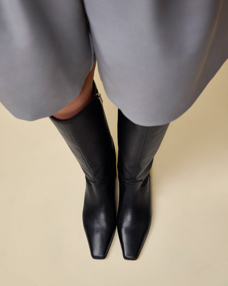A person in a knee-length gray skirt and Vince Camuto Pavla Narrow Calf Knee High Boots stands on a light beige floor, viewed from above.