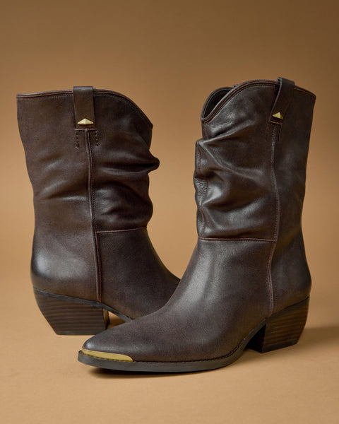 The Vince Camuto Pezzra Western Bootie is a mid-calf, dark brown leather cowboy boot with a slouchy design, stacked heels, and pointed toes, shown against a plain brown background.