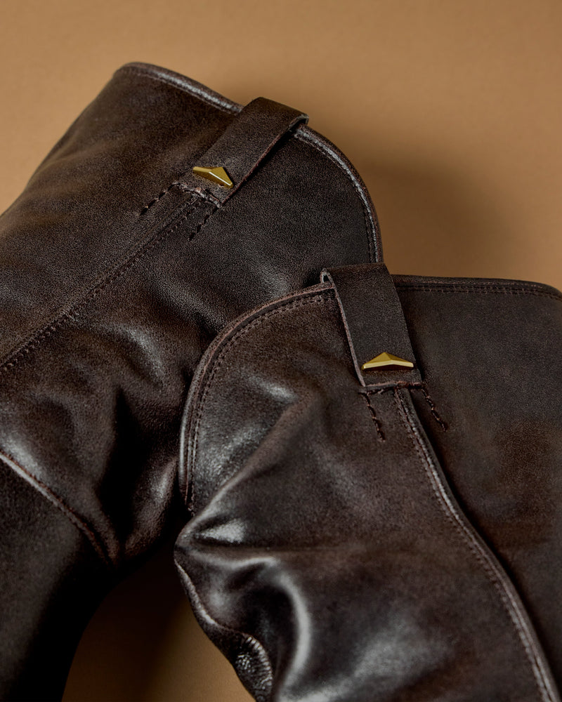 Close-up of the Vince Camuto Pezzra Western Bootie, featuring brown leather, visible stitching, and gold-toned pull tab accents, set against a tan background.