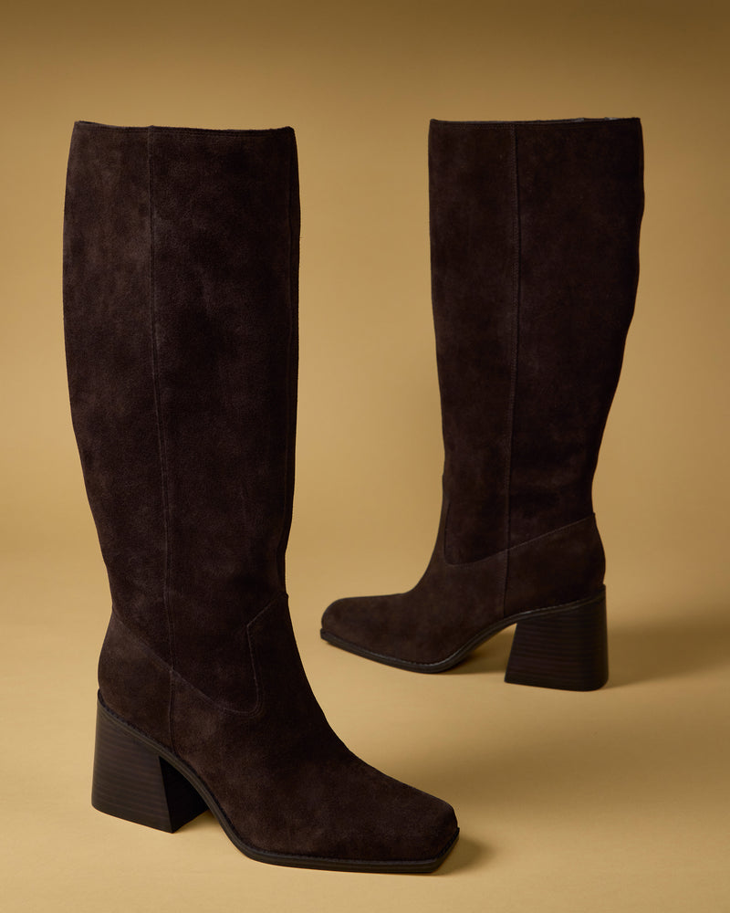 The Vince Camuto Sangeti Narrow Calf Boot, a pair of tall dark brown suede boots with block heels and square toes, is shown on a beige background—one upright and the other angled slightly to the side.