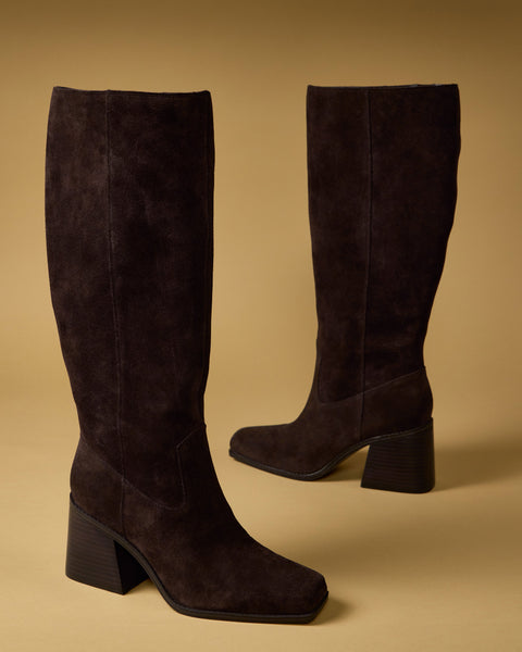 A pair of Vince Camuto Sangeti Boots in dark brown suede with block heels, displayed on a light tan surface against a matching background.