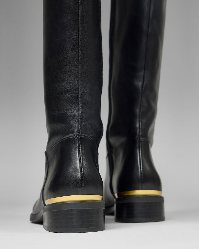 Close-up of the back of Vince Camuto Bercla Wide Calf Knee High Riding Boots in black leather, featuring low stacked heels with gold metallic accents, set against a plain gray background.