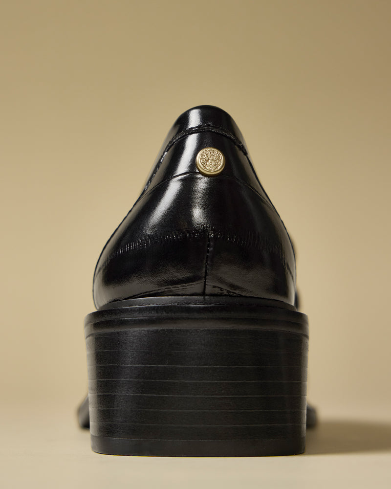 A close-up rear view of the Vince Camuto Eshnie Block Heel Loafer in shiny black leather with a stacked block heel and a gold emblem on the heel counter, shown against a beige background.