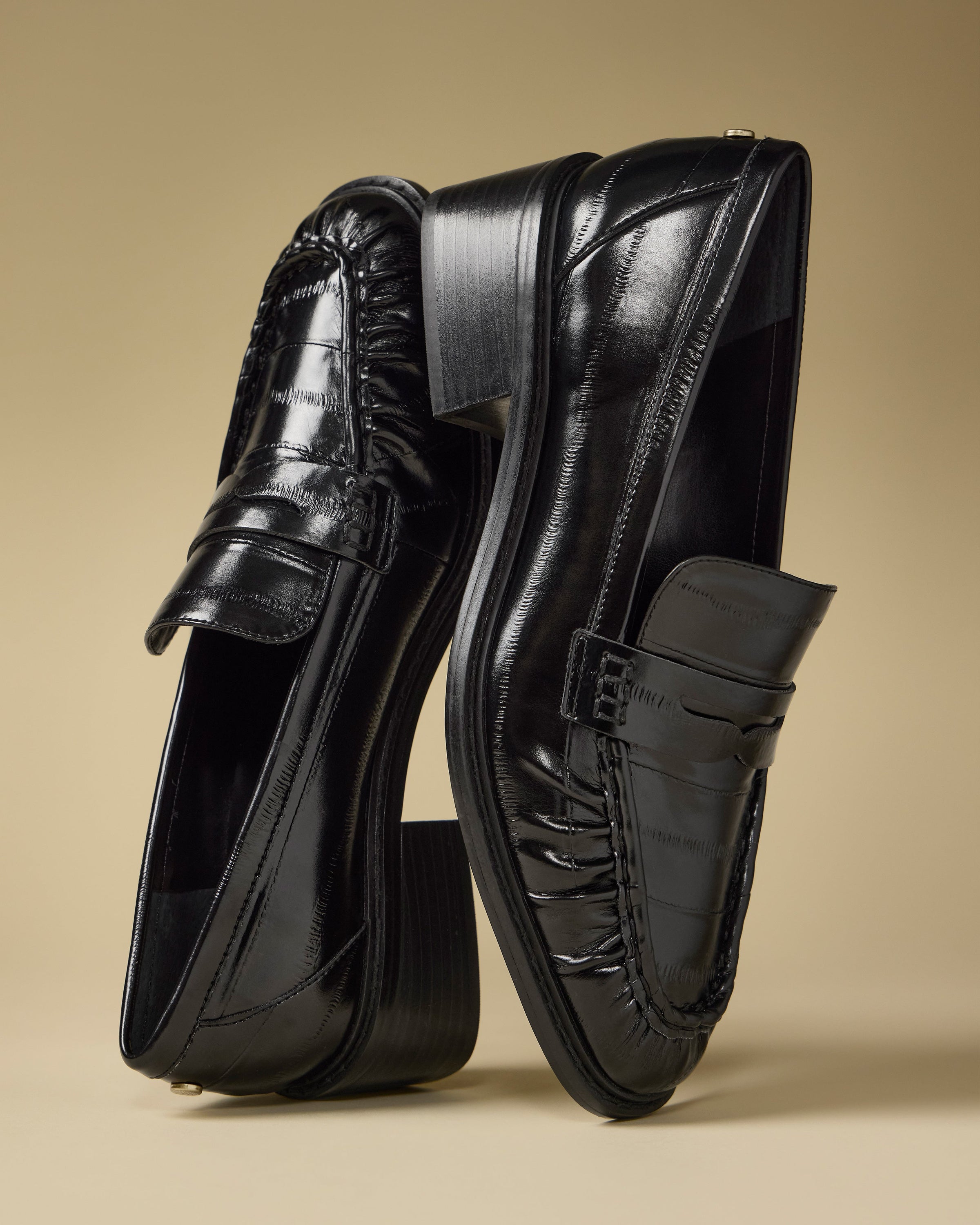 A pair of Vince Camuto Eshnie Block Heel Loafers in shiny black—one upright, the other leaning against it—are displayed on a plain beige background.