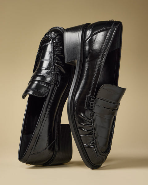 A pair of Vince Camuto Eshnie Block Heel Loafers in shiny black—one upright, the other leaning against it—are displayed on a plain beige background.