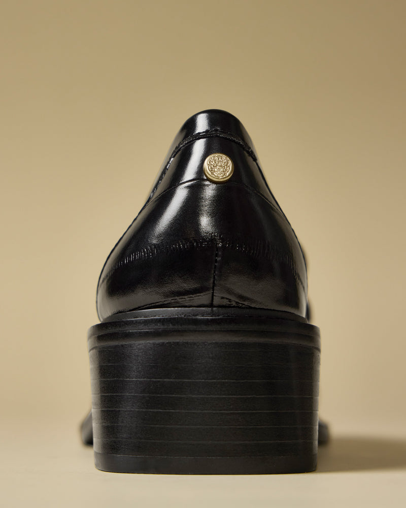 A close-up shows the back of the Vince Camuto Eshnie Block Heel Loafer in shiny black with a stacked heel and small round gold emblem near the collar, set against a light beige background.
