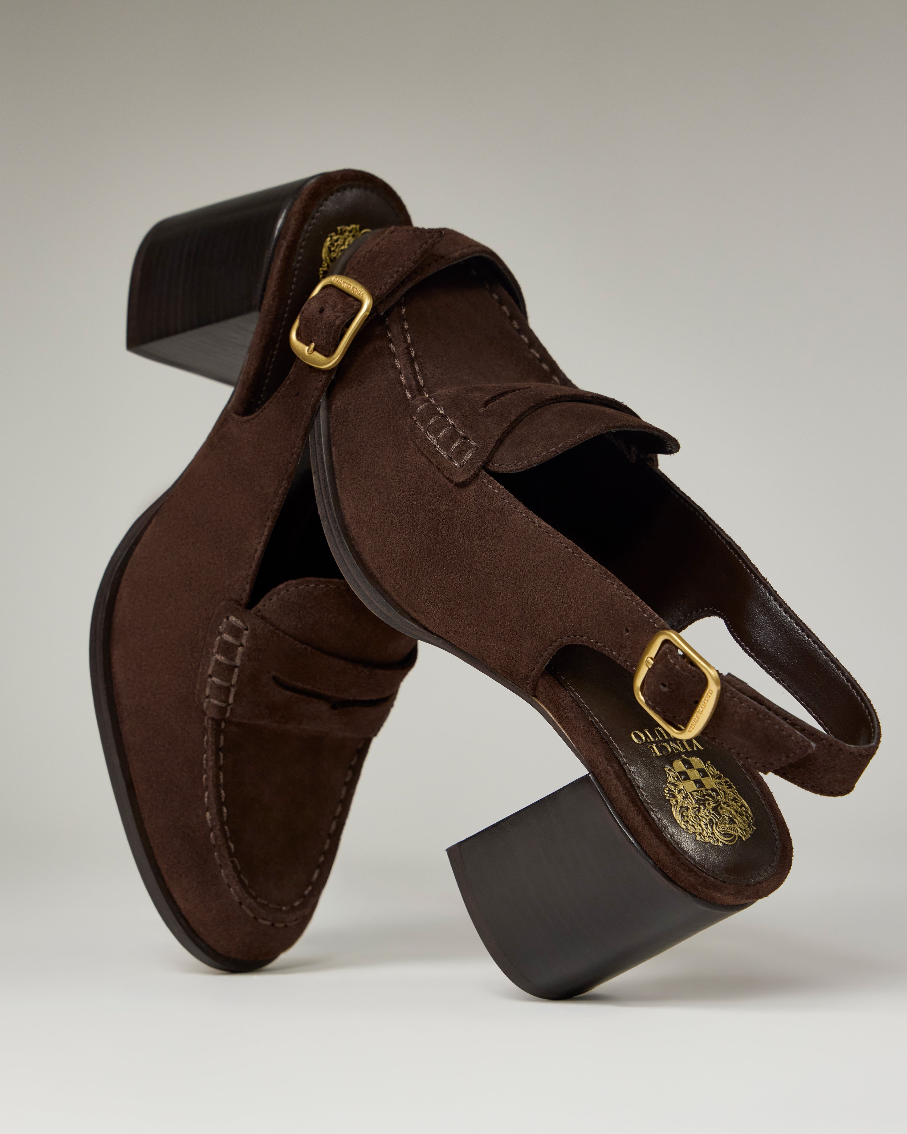 The Vince Camuto Grinie Loafer features brown suede, chunky block heels, gold buckles, a Penny Keeper strap detail, visible stitching, and dark wooden heels, styled artfully on a neutral backdrop.