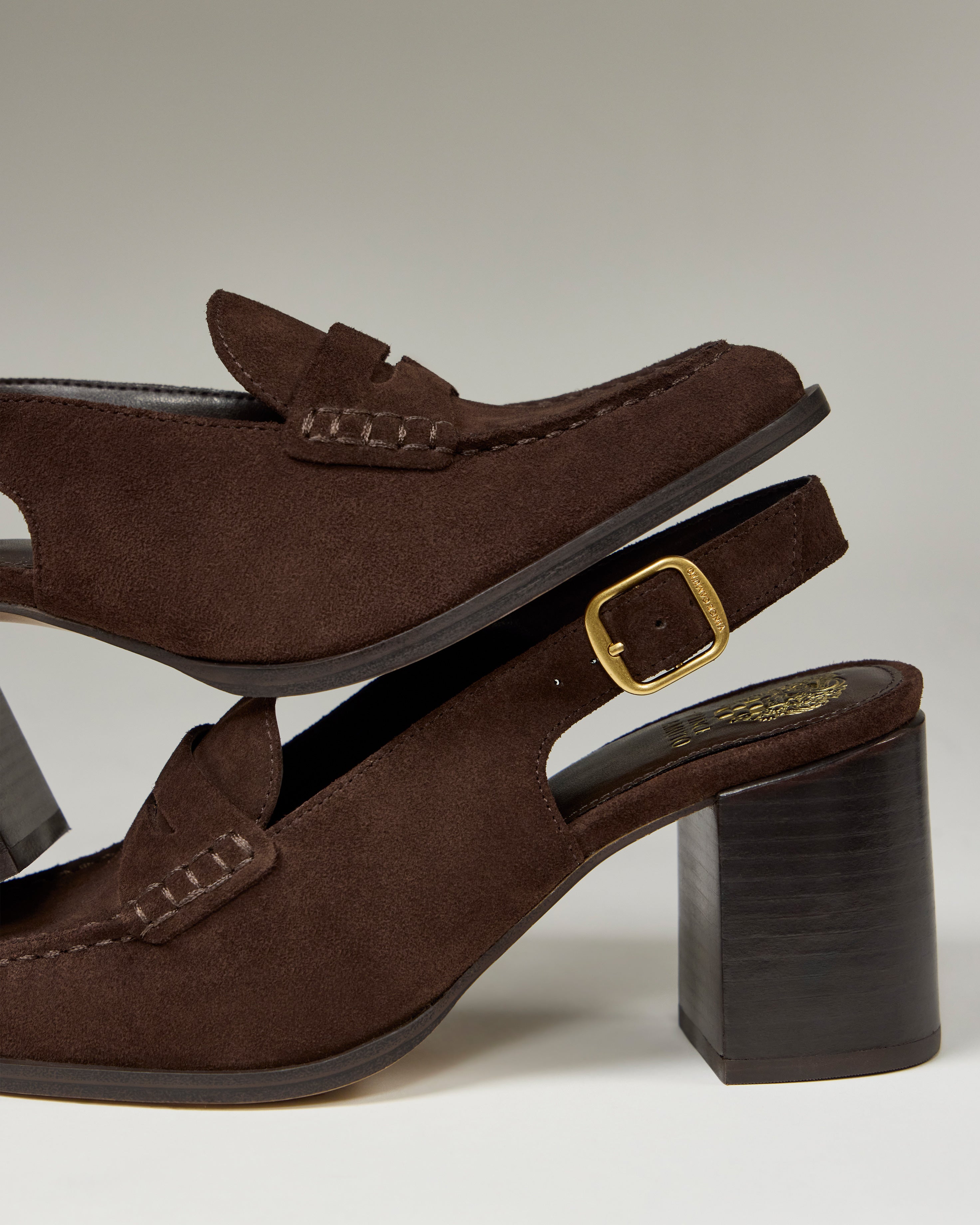 Two Vince Camuto Grinie Loafers are stacked, both in brown suede with chunky block heels. The bottom shoe has a gold buckle and visible stitching, while both display classic penny loafer style with Penny Keeper straps.