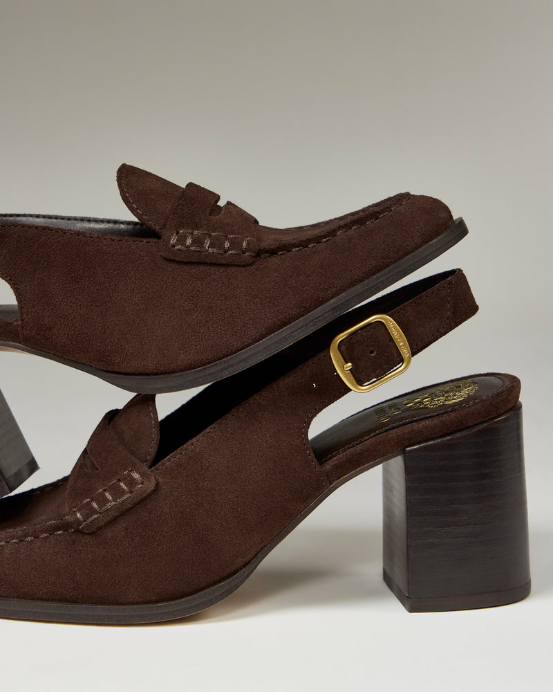 Two Vince Camuto Grinie Loafers are stacked, both in brown suede with chunky block heels. The bottom shoe has a gold buckle and visible stitching, while both display classic penny loafer style with Penny Keeper straps.