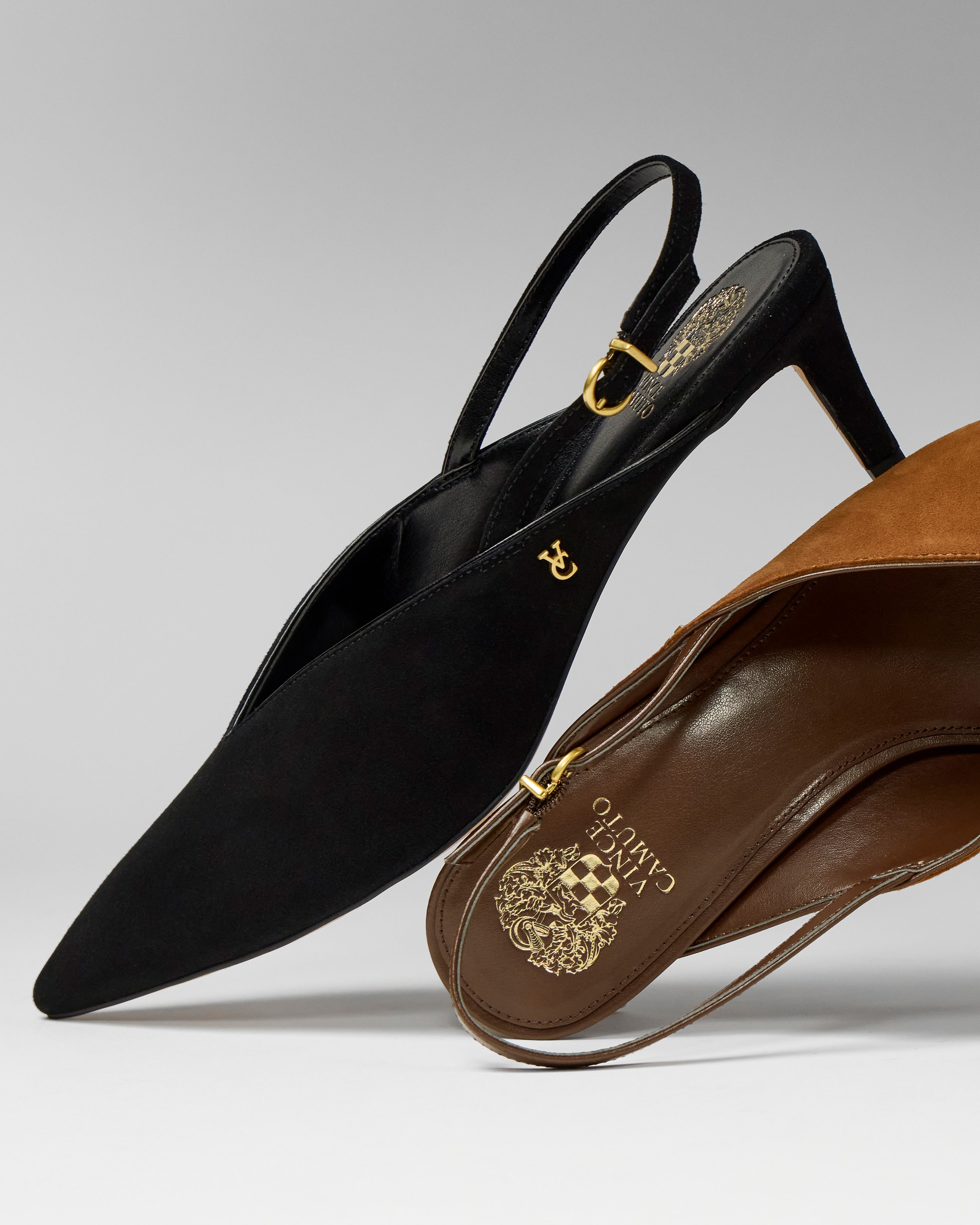 A close-up of two Vince Camuto Kylien Kitten Heel Pumps: one in black suede with a gold buckle and logo, the other in brown leather with gold crest branding—both feature timeless mid-height pointed heels.