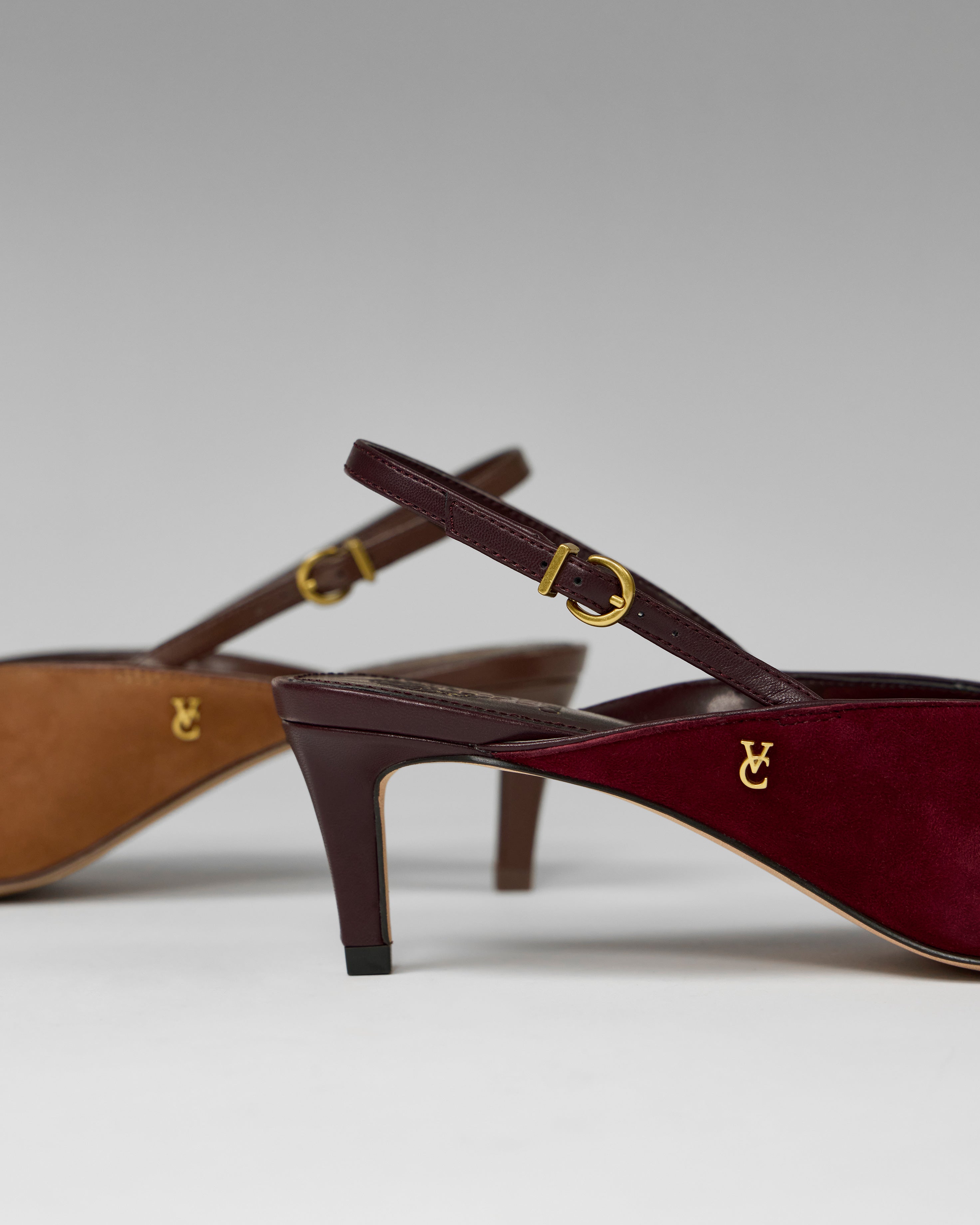 Close-up of two Vince Camuto Kylien Kitten Heel Pumps, one burgundy and one tan, both featuring gold buckles and a small gold logo by the kitten heel, displayed against a neutral background.