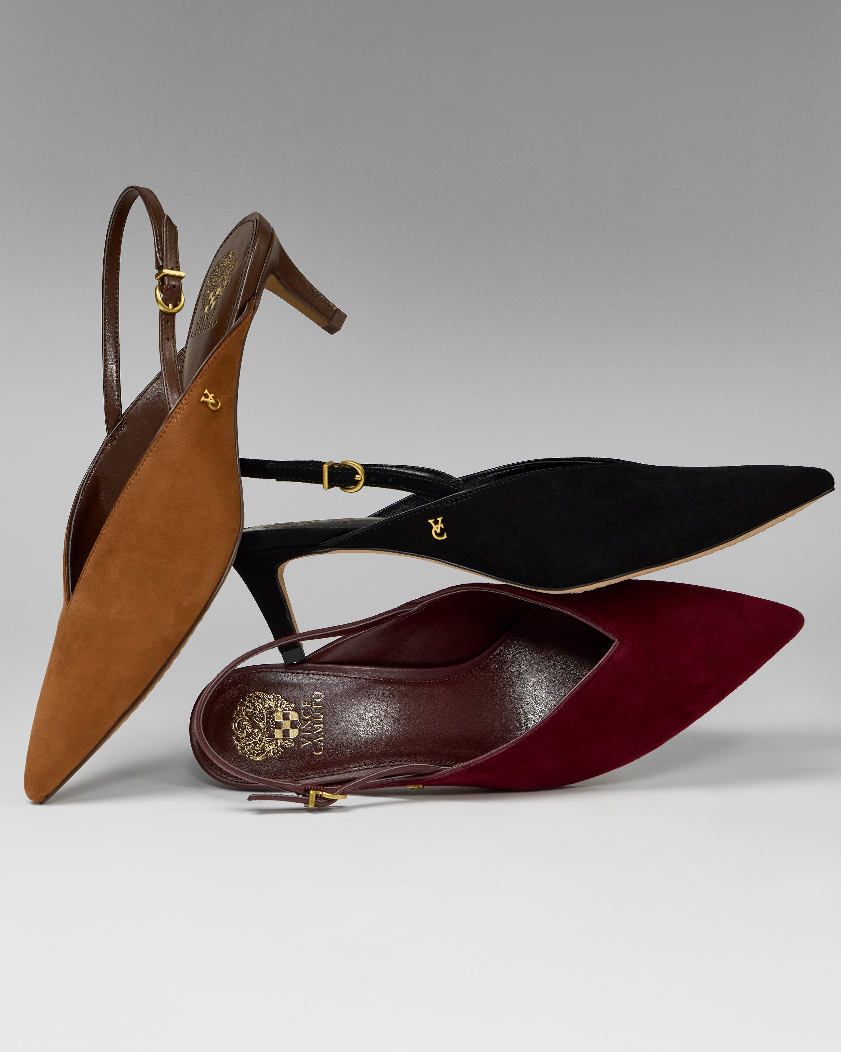 Three Vince Camuto Kylien Kitten Heel Pumps in brown, black, and burgundy suede are stacked on a gray background. Each pointed-toe slingback features a gold buckle accent and small gold emblem on the side.