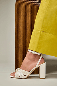 A person wearing a yellow skirt and white woven-strap block heel sandals stands next to a textured brown surface.