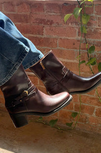 A pair of dark brown leather ankle boots with side buckles, worn with blue jeans, set against a brick wall background with green ivy.