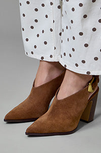 A close-up of a person wearing brown suede pointed-toe heeled mule boots with a gold accent heel puller, paired with white pants featuring a black polka dot pattern.