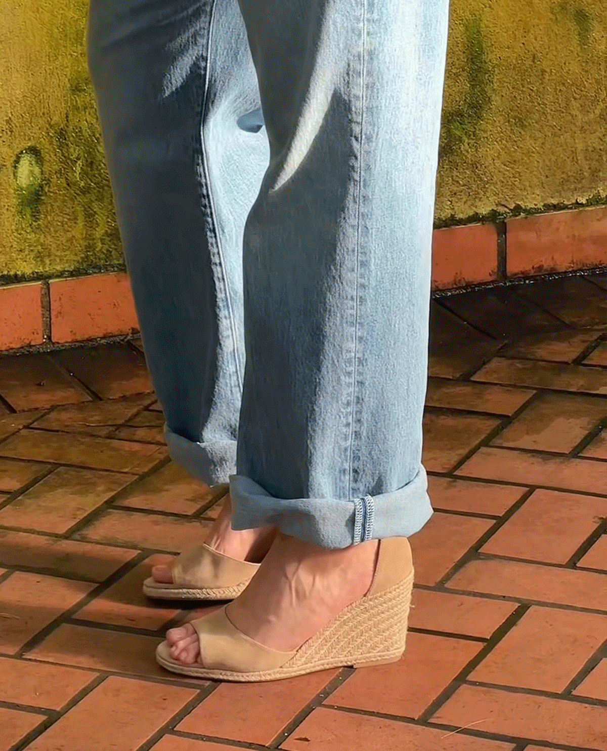 A person standing on a brick floor wearing light blue cuffed jeans and beige wedge sandals. A yellowish, weathered wall is in the background.