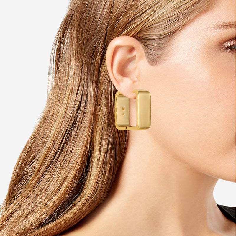 A close-up shows a woman with straight, light brown hair tucked behind her ear, wearing Vince Camuto Brass Rectangle Hoop Earrings that feature a modern geometric design and highlight their bold, gold-tone elegance against her fair skin.