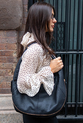 A woman with long dark hair and glasses stands sideways against a brick wall, wearing a sheer white blouse with black polka dots and carrying a large black shoulder bag.