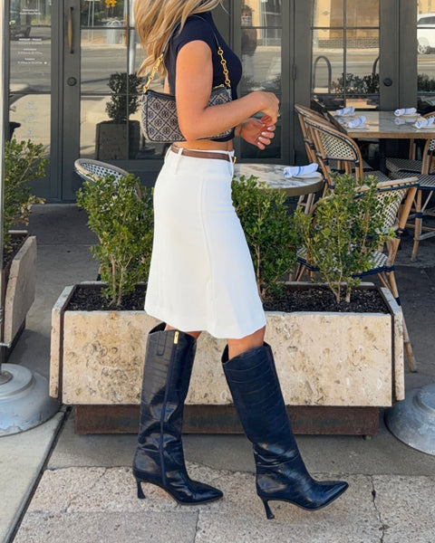 A woman in a black top, white skirt, and the Vince Camuto Kenlea Knee High Boot walks outside a café with outdoor seating, carrying a patterned shoulder bag.