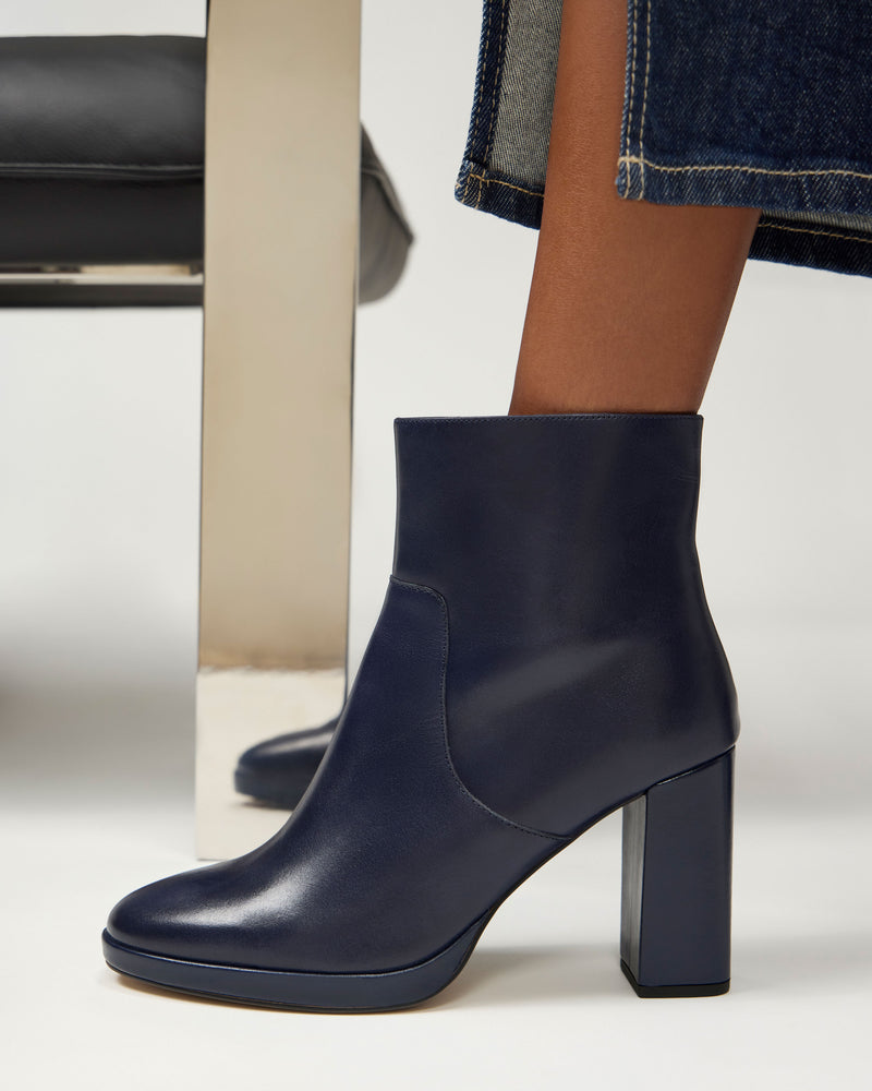 A close-up of a person wearing the Laylei Bootie by Vince Camuto in navy blue, showcasing its stylish block heel. The booties are paired with dark blue jeans with frayed hems, and the scene includes a metal chair leg.