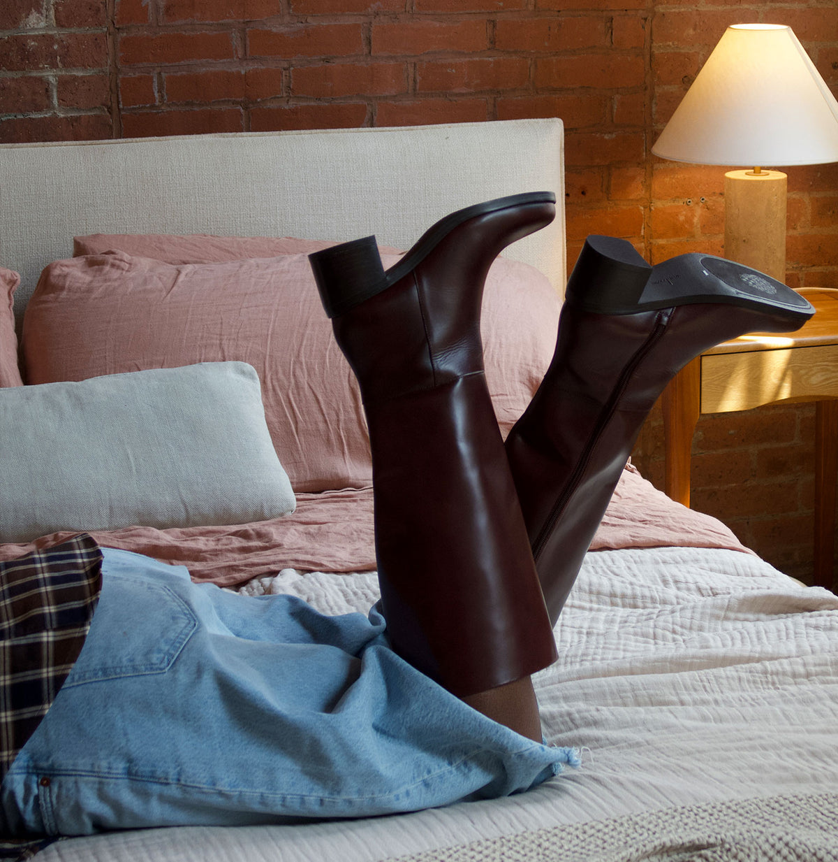 A person reclining on a bed with their legs raised, wearing a pair of dark brown leather over-the-knee boots. The bedroom setting features a brick wall, a lit bedside lamp, and pink bedding.
