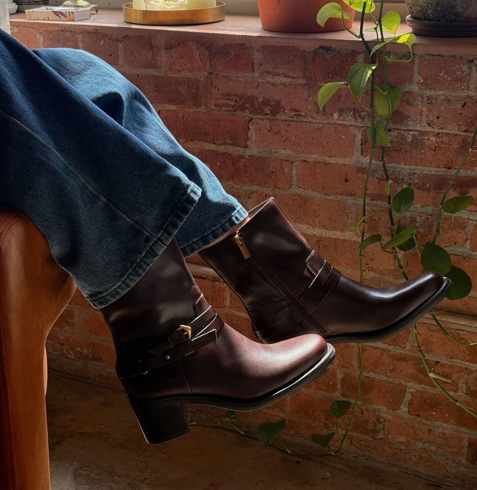 A person wearing dark brown leather ankle boots with side buckles and blue jeans, sitting on a leather chair in front of a brick wall with a hanging plant.