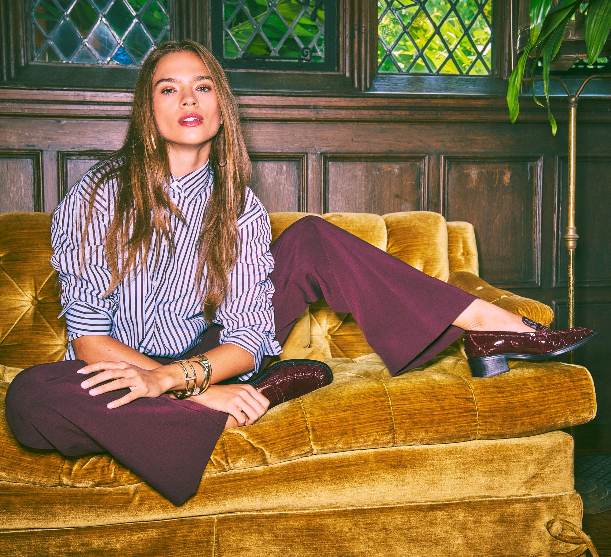 A woman sits on a mustard-yellow velvet sofa in a rustic room with dark wood paneling and diamond-patterned windows. She is wearing a lavender and white vertically striped button-down shirt, dark burgundy wide-leg trousers, and dark burgundy patent leather shoes.