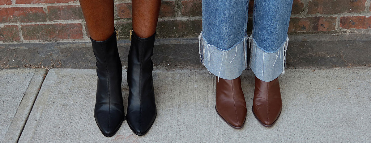 Two models standing side by side wearing pointed-toe ankle boots, one in black leather and one in brown leather, paired with rolled-up jeans against a brick wall and concrete sidewalk.