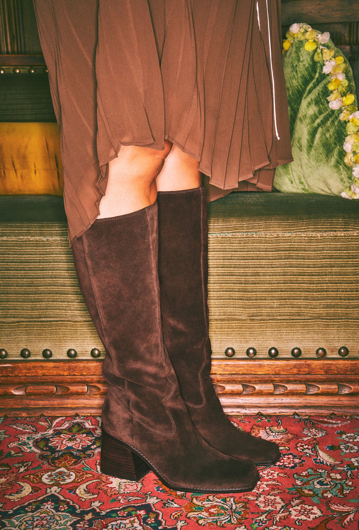 A person wearing tall dark brown suede knee-high boots with a block heel, paired with a brown pleated skirt, standing on a patterned red rug in front of a vintage green bench.
