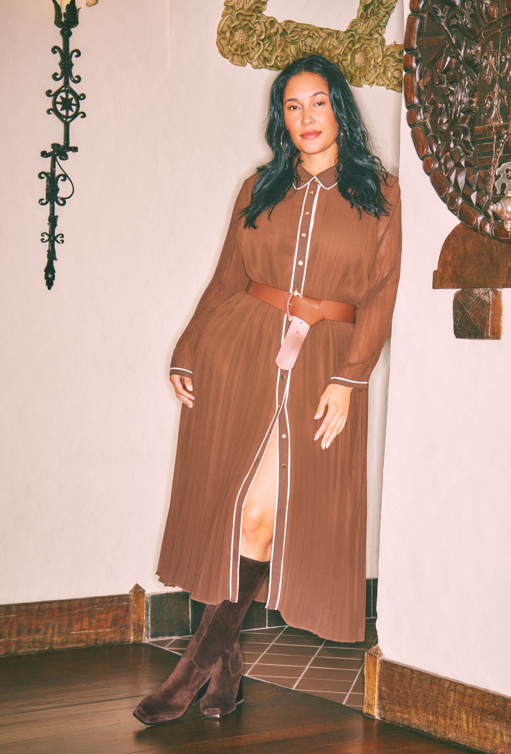 A woman models a brown long-sleeve midi shirt dress with white piping details, a white collar, and a matching brown belt. She is wearing dark brown heeled boots in an indoor setting with a tiled floor and decorative wall art.