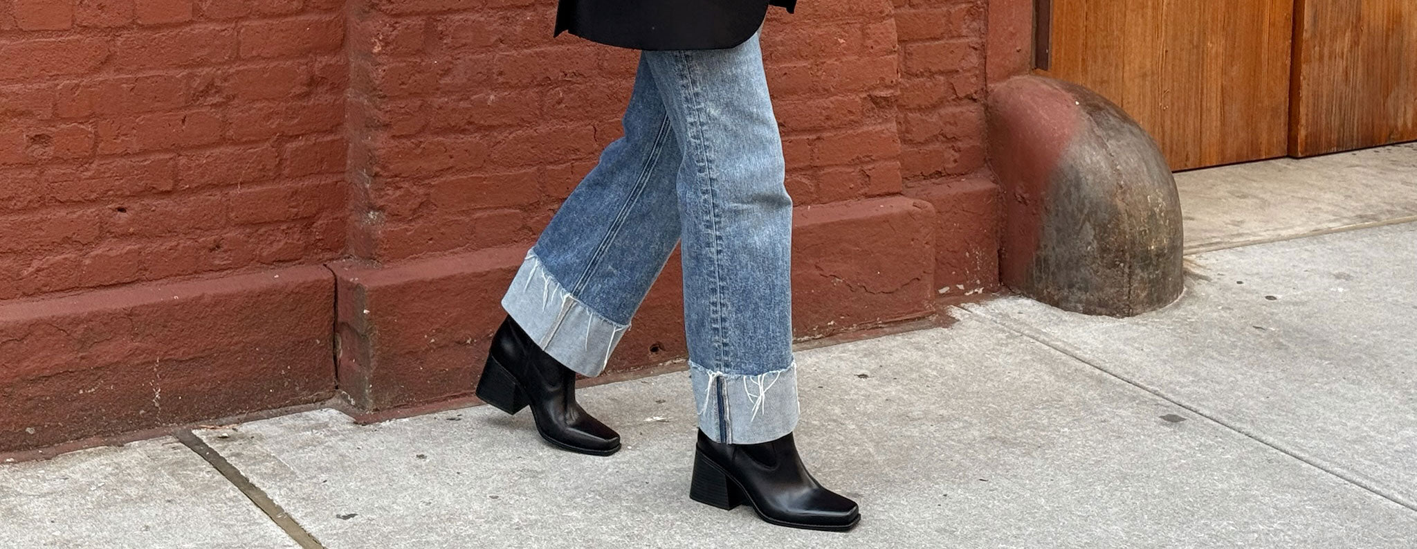 A person walking on a sidewalk wearing light blue wide-leg jeans with turned-up cuffs, black ankle boots, and a black top, set against a red brick wall and concrete ground.