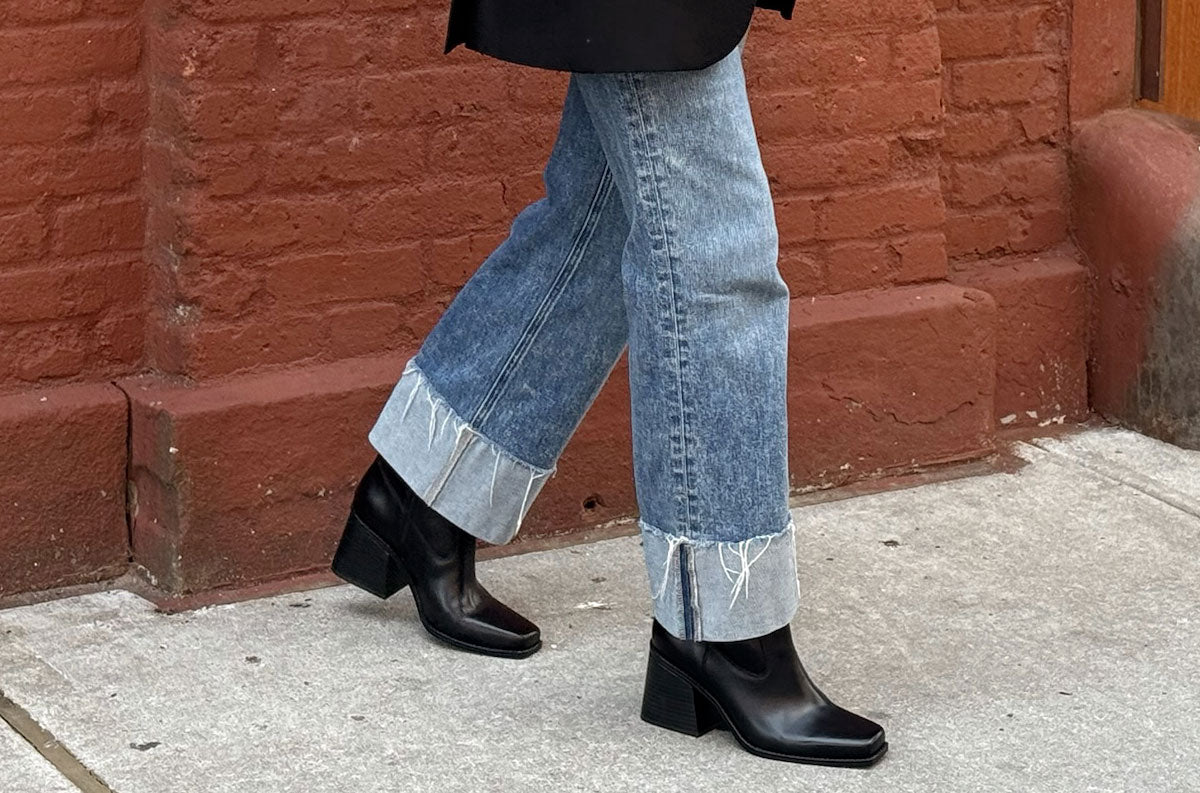 A person wearing light blue cuffed jeans and black ankle boots walking on a sidewalk in front of a red brick wall.