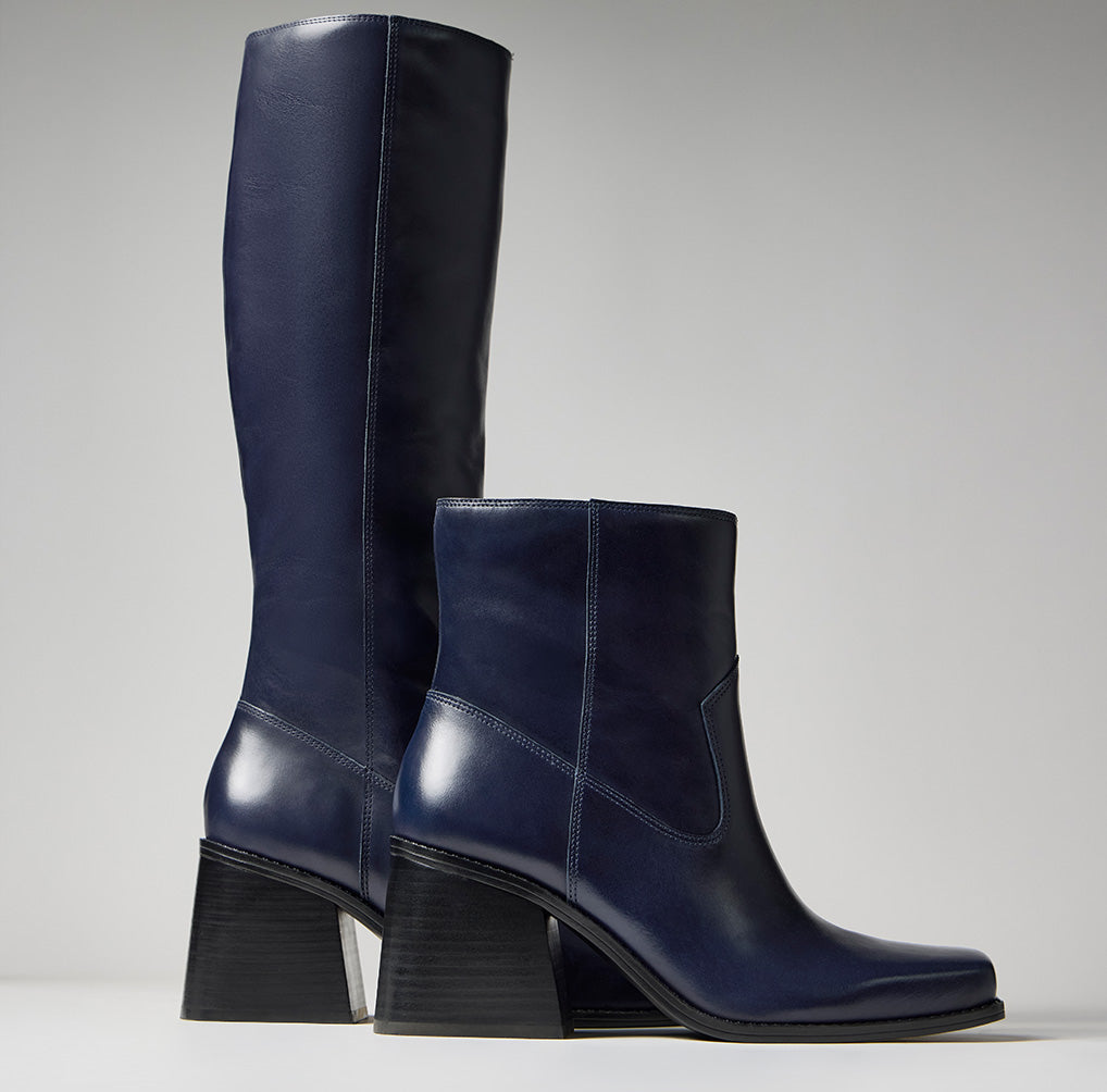 A pair of navy blue leather knee-high boots with chunky black block heels, shown in a studio setting against a light gray background.