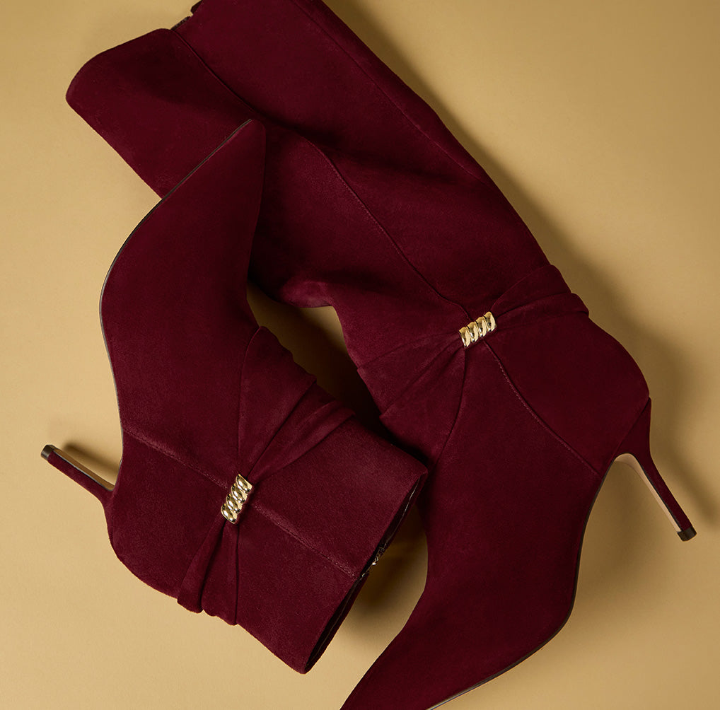 A pair of deep burgundy suede knee-high boots with pointed toes and stiletto heels, decorated with gold-tone chain-link embellishments near the ankles, displayed on a solid beige background.